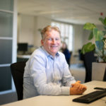 Business professional sitting at a desk, smiling confidently in a modern office environment.